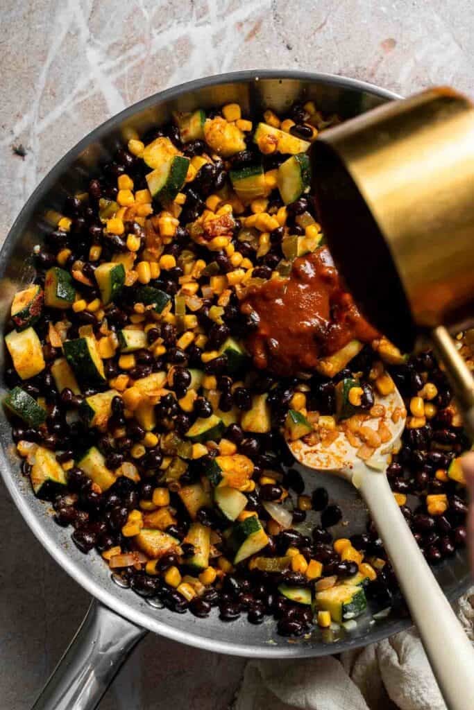Enchilada sauce being poured into a skillet of vegetable filling