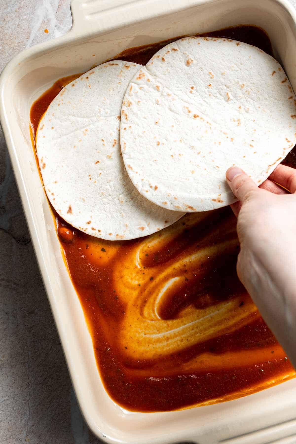 Tortillas being layered over enchilada sauce in a casserole dish
