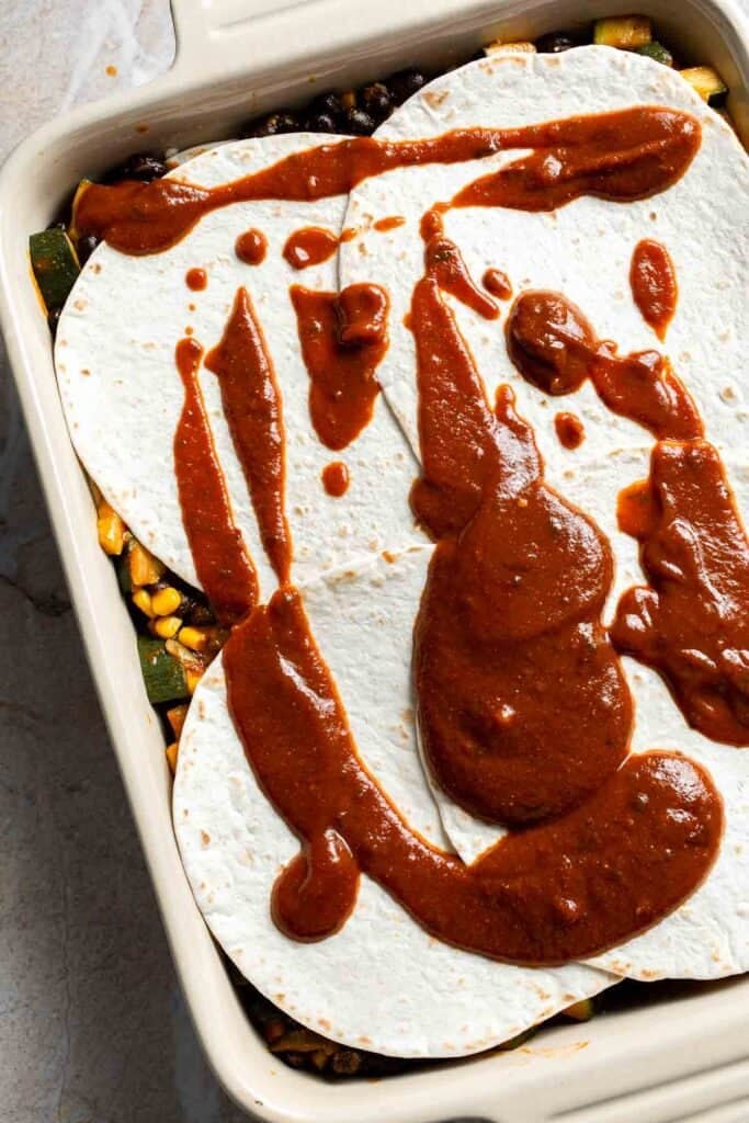 Enchilada sauce being drizzled over the final tortilla layer in a casserole dish
