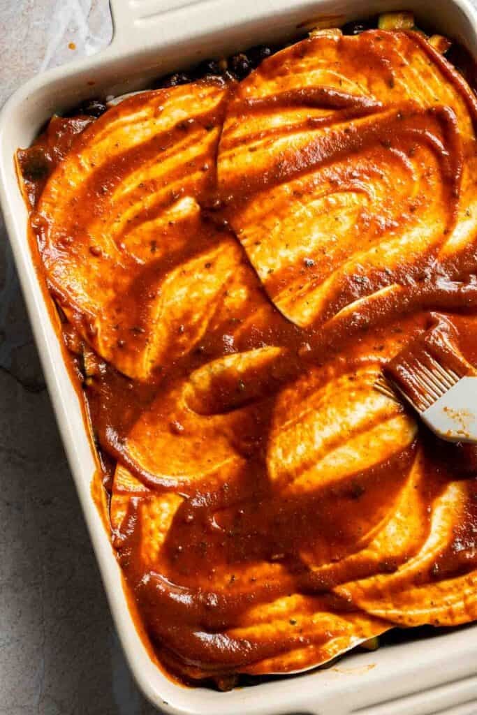 Enchilada sauce spread evenly over the top tortilla layer in a casserole dish