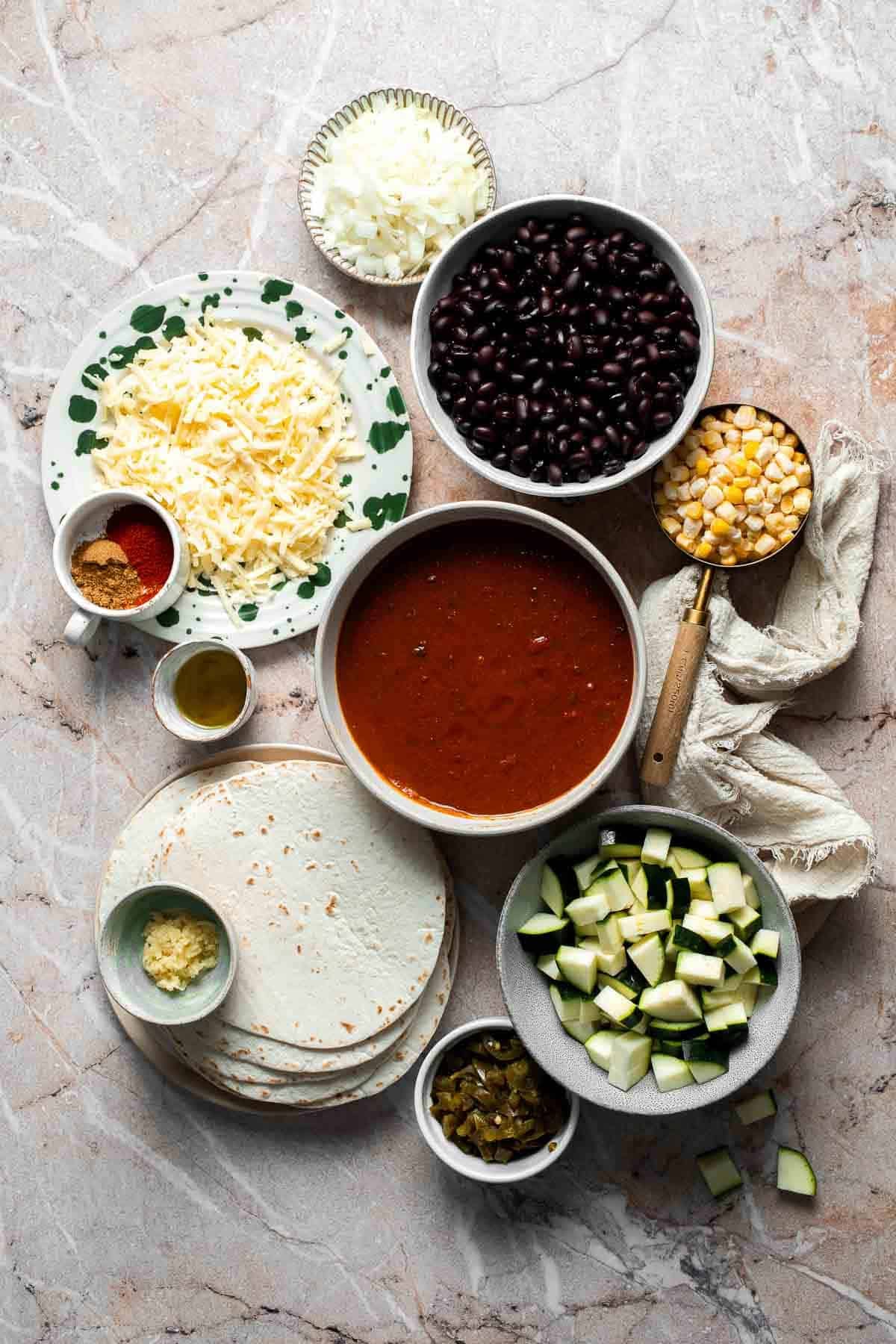 Ingredients for vegetarian enchilada casserole including black beans, cheese, tortillas, and enchilada sauce arranged on a counter