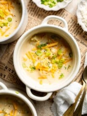 Bowl of cheesy cauliflower soup topped with shredded cheddar and chopped green onions, shown from above with extra toppings on the side.