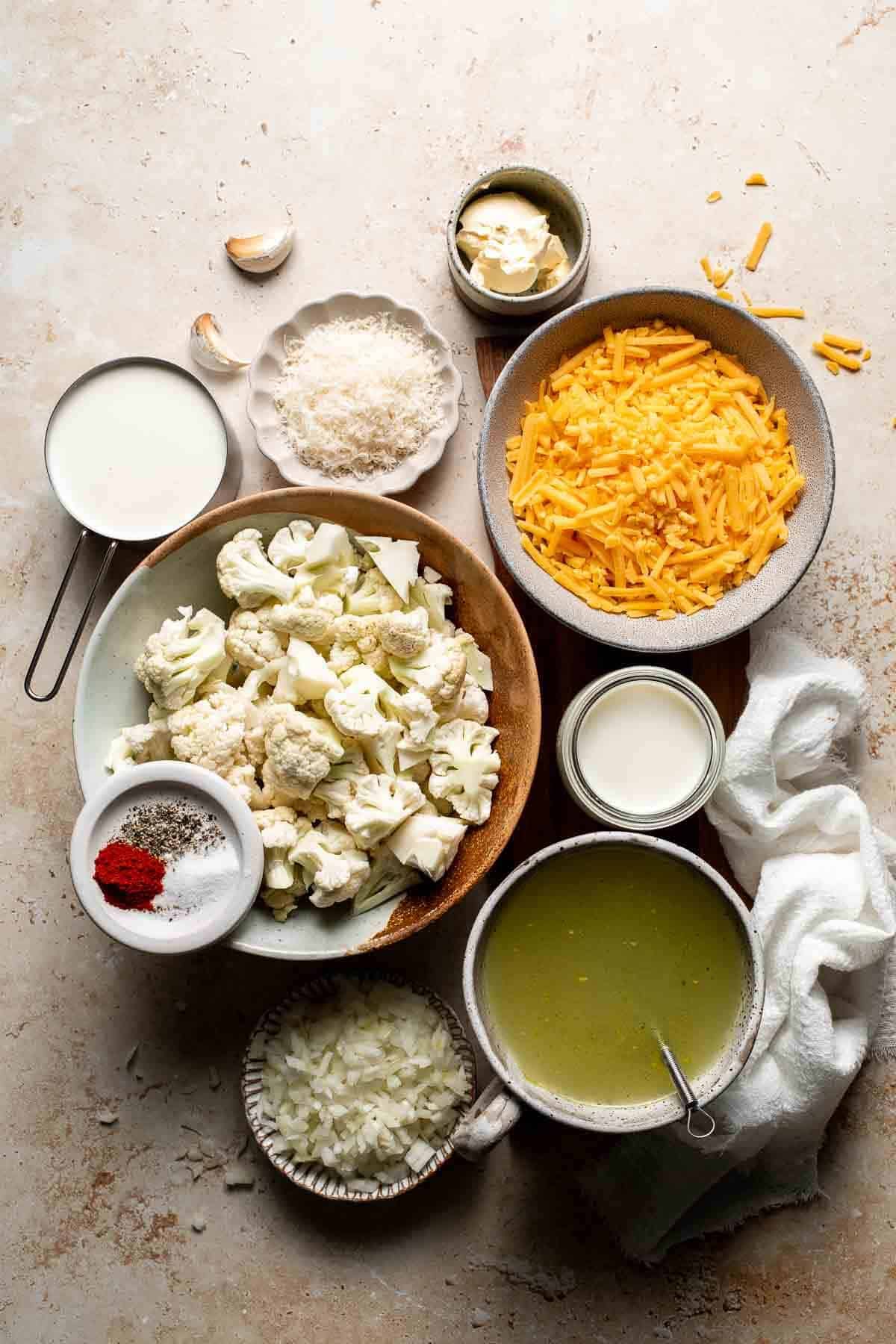 Ingredients for cheesy cauliflower soup, including cauliflower florets, shredded cheddar, milk, vegetable broth, garlic, and paprika, arranged on a light surface.