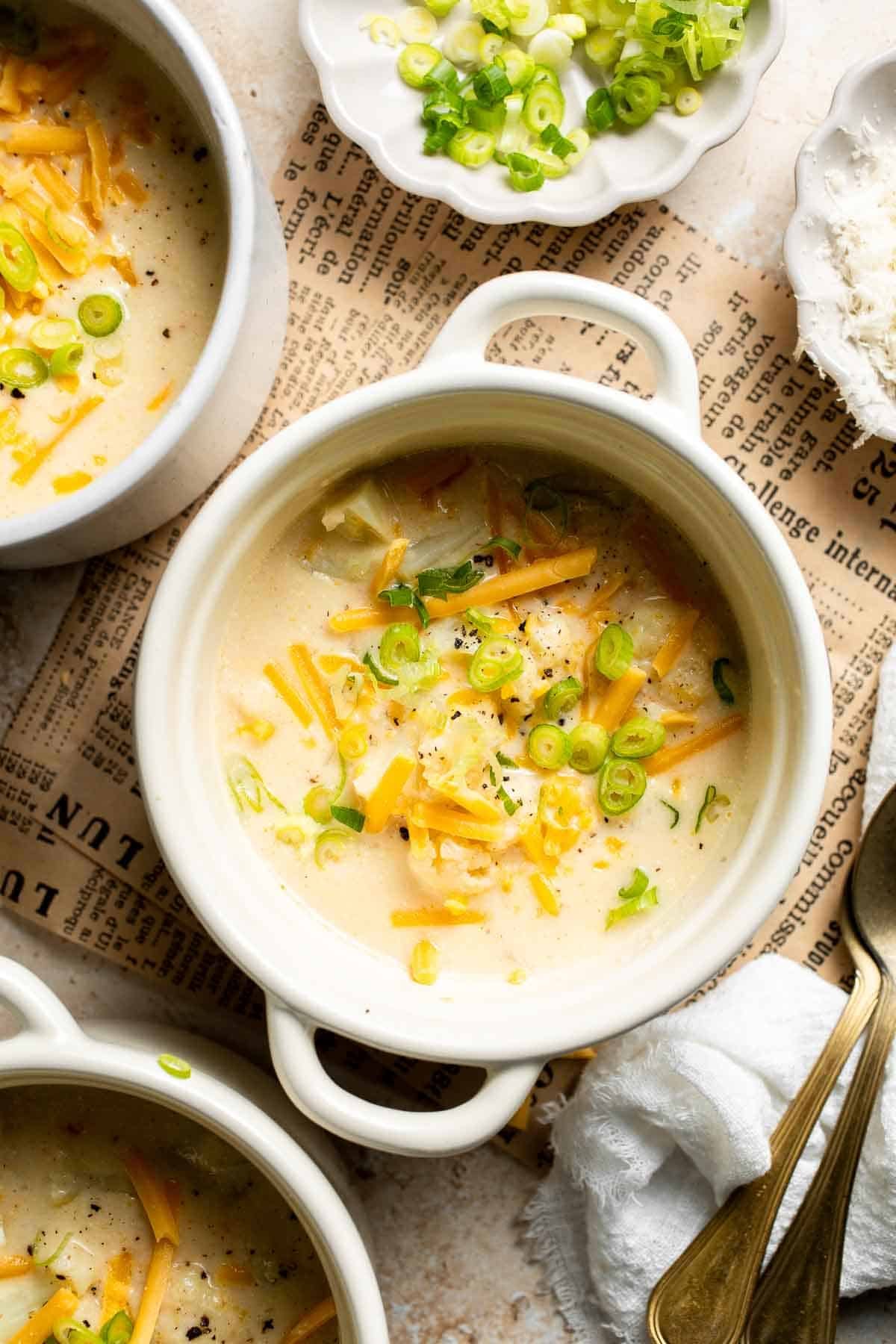 Bowl of cheesy cauliflower soup topped with shredded cheddar and chopped green onions, shown from above with extra toppings on the side.