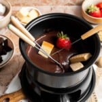 Chocolate fondue in a black fondue pot with a strawberry and cake cube on fondue forks, surrounded by dippers.