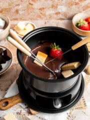 Chocolate fondue in a black fondue pot with a strawberry and cake cube on fondue forks, surrounded by dippers.