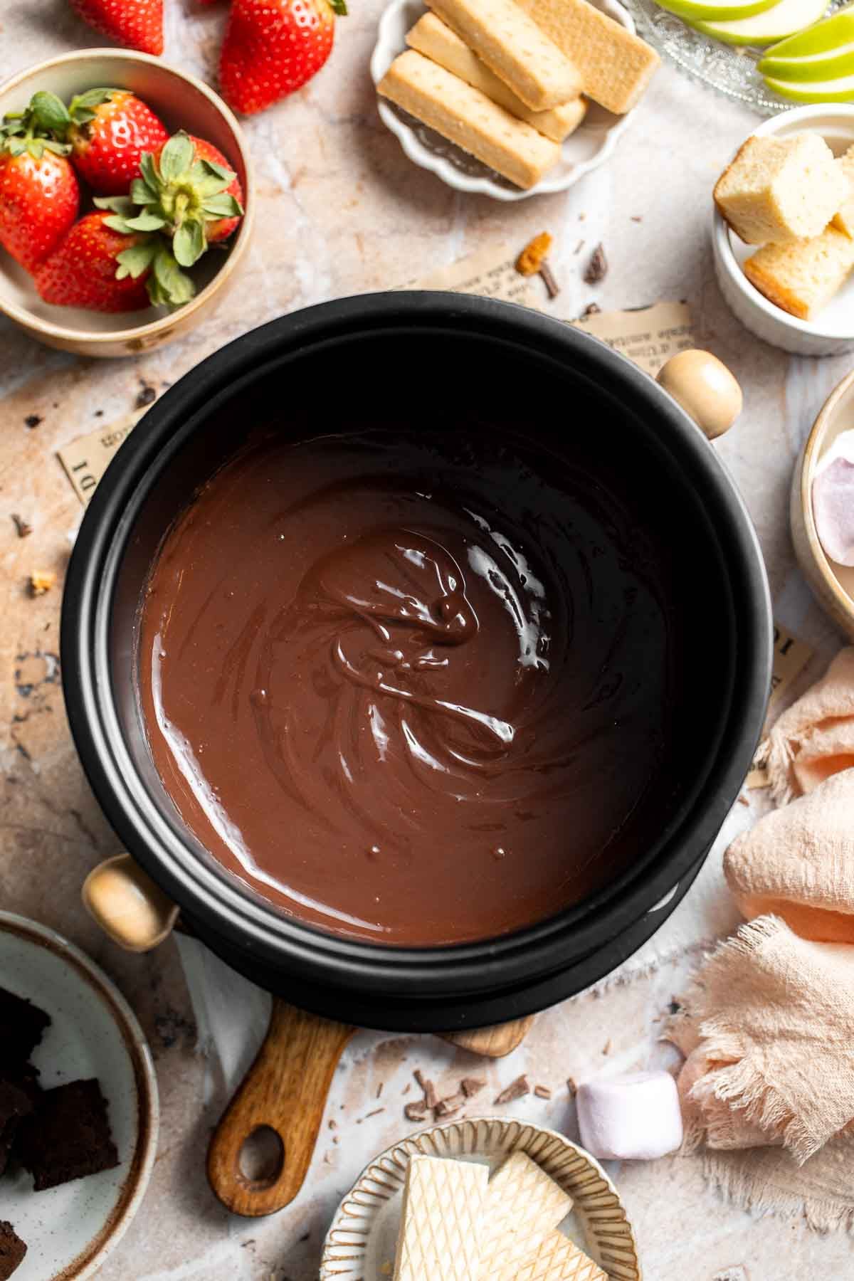 Overhead view of silky chocolate fondue in a black fondue pot, surrounded by strawberries, apple slices, cake, cookies, and marshmallows.