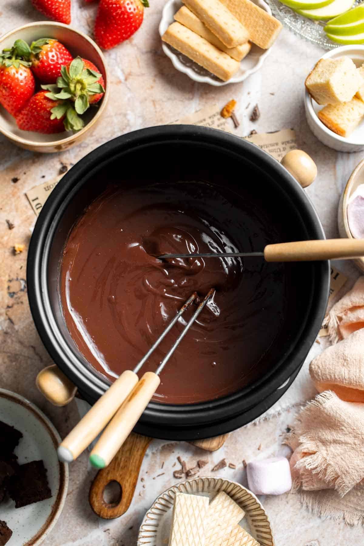 Fondue forks resting in a pot of melted chocolate fondue, with dippers around the pot.