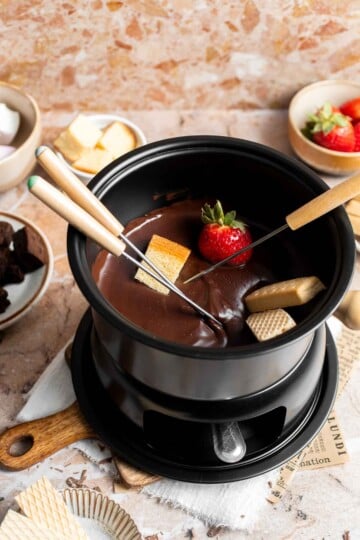 Chocolate fondue in a black fondue pot with a strawberry and cake cube on fondue forks, surrounded by dippers.
