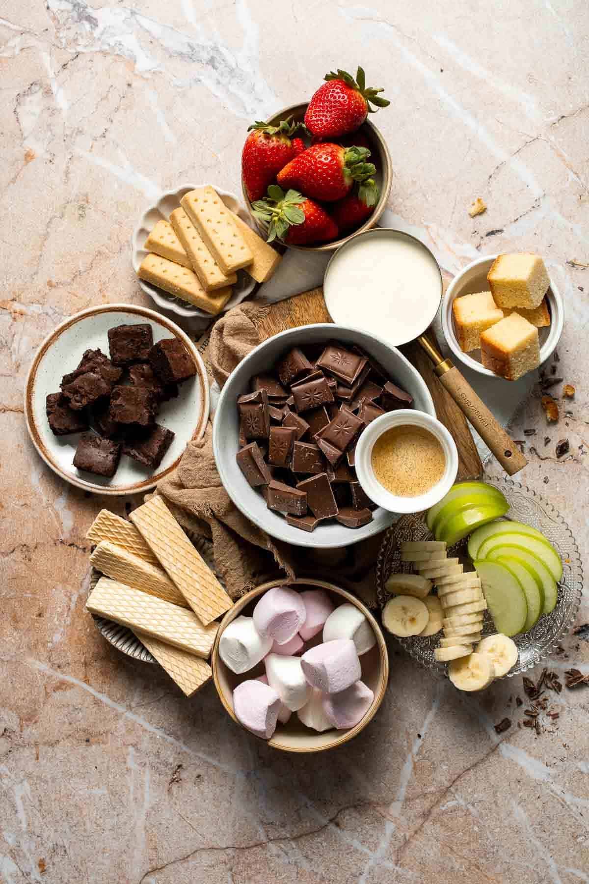 Chocolate fondue ingredients and dippers, including chopped chocolate, heavy cream, strawberries, apple and banana slices, marshmallows, shortbread, and wafer cookies, arranged on a marble surface.