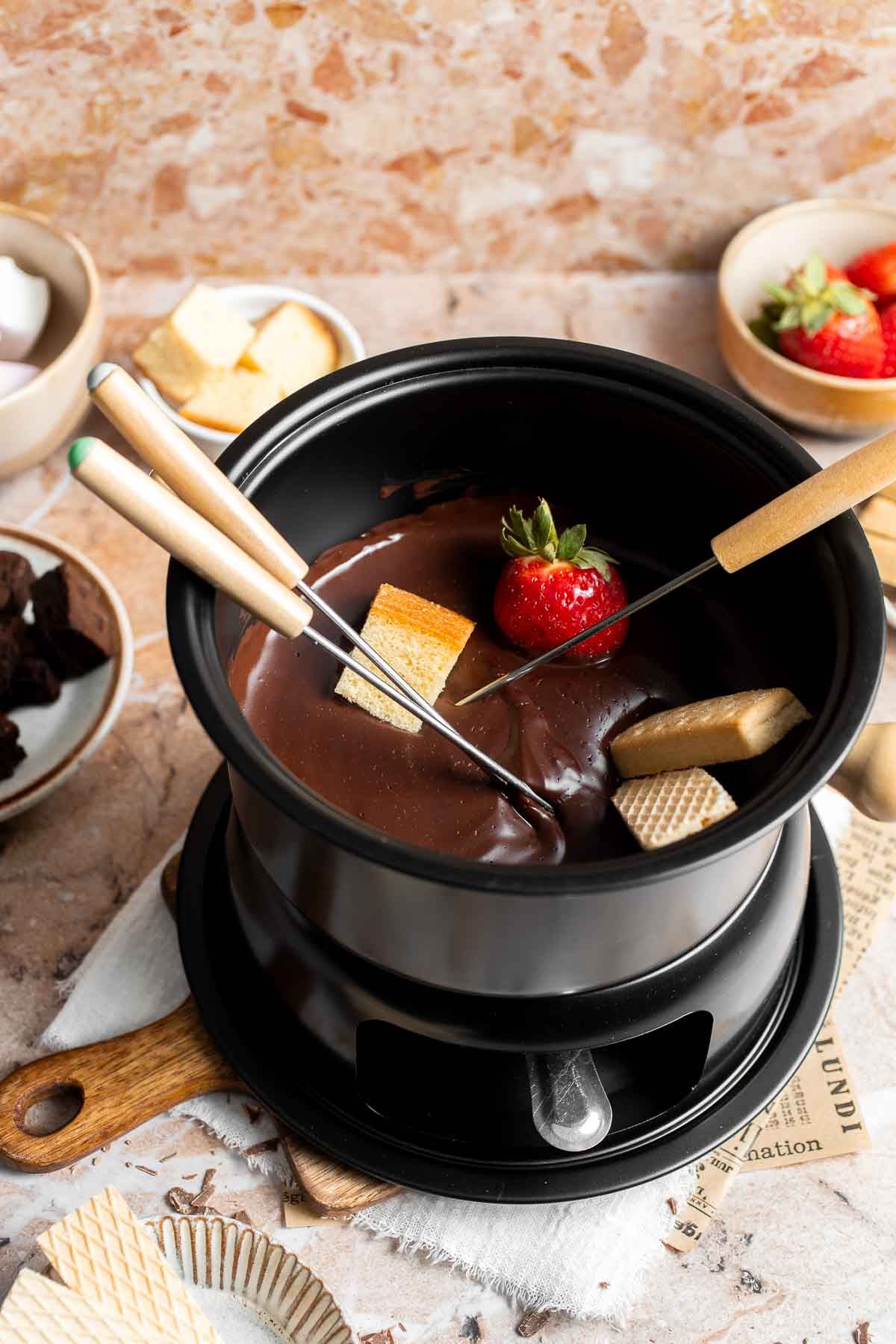 Chocolate fondue in a black fondue pot with a strawberry and cake cube on fondue forks, surrounded by dippers.