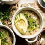 Creamy white bean chili in white bowls topped with fanned avocado slices, lime wedges, sliced jalapeños, and fresh cilantro, on a newspaper-lined board.