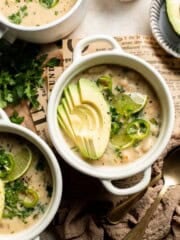Creamy white bean chili in white bowls topped with fanned avocado slices, lime wedges, sliced jalapeños, and fresh cilantro, on a newspaper-lined board.