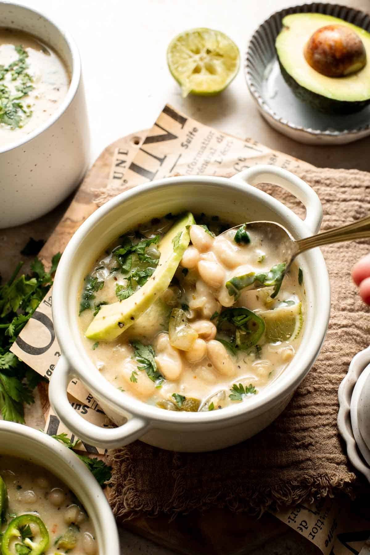 Creamy white bean chili in a white bowl with whole cannellini beans, avocado, cilantro, and jalapeño, with a gold spoon.