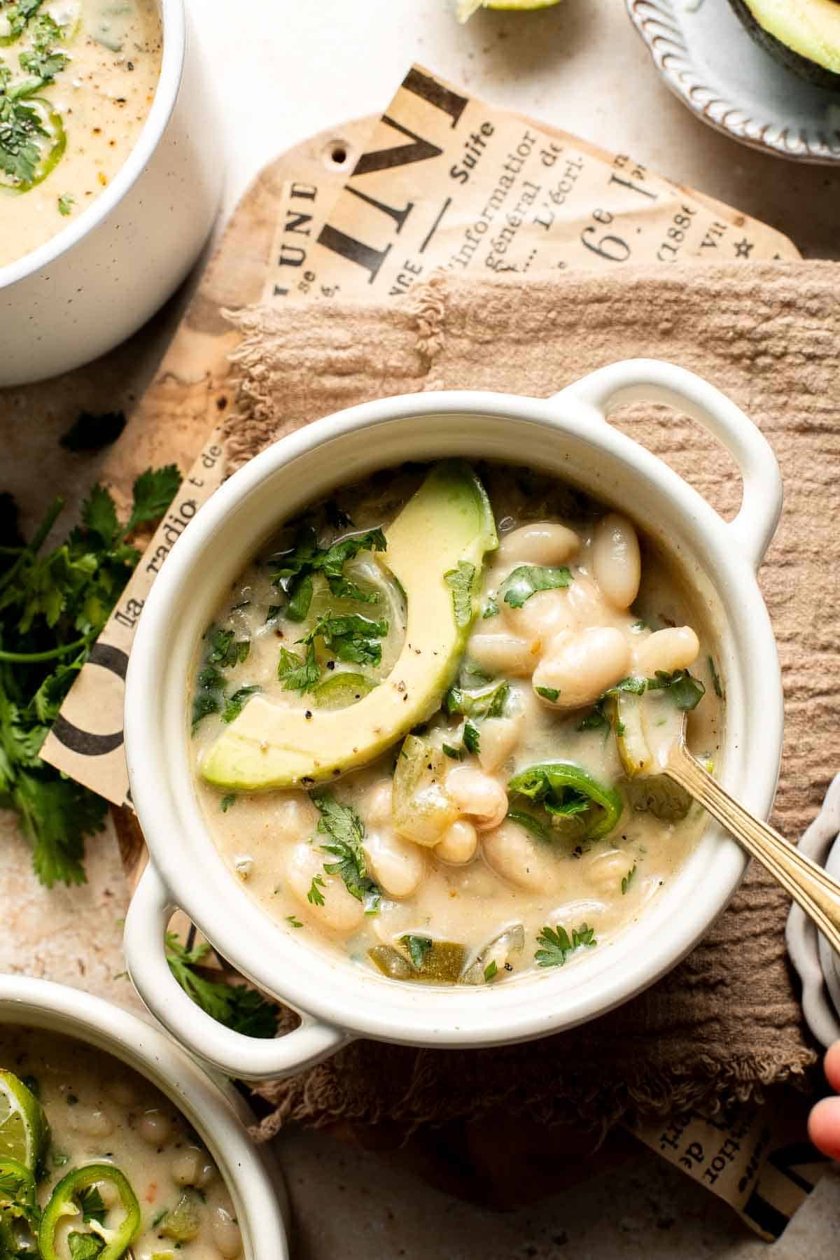 Overhead view of creamy white bean chili in a white bowl with an avocado slice, cilantro, and jalapeño, on a linen cloth and newspaper print.