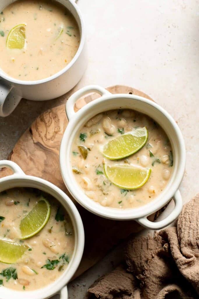 Bowls of creamy white bean chili topped with lime slices, on a wooden board with a linen cloth.