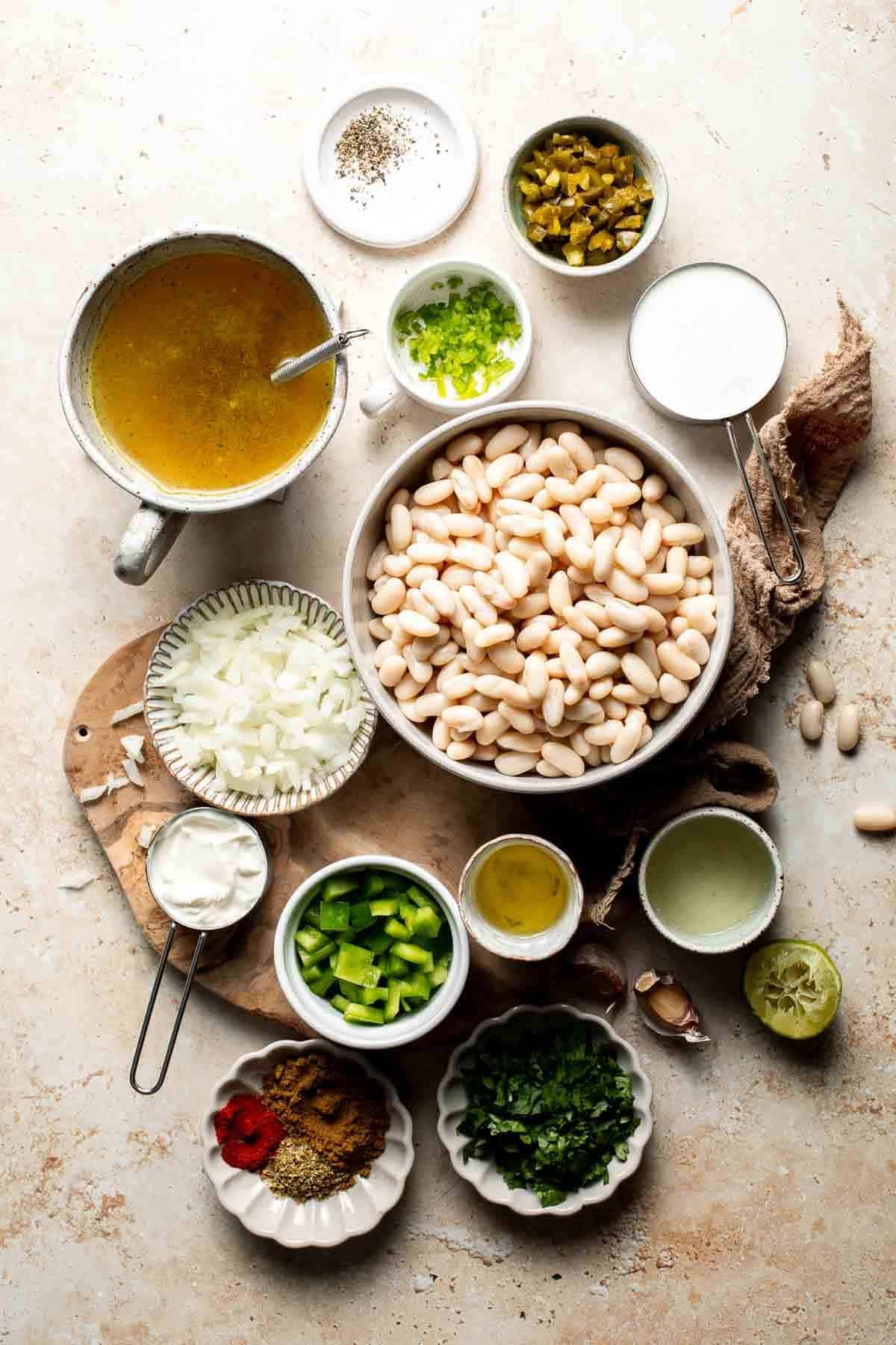 Ingredients for creamy white bean chili, including vegetable broth, cannellini beans, diced onion, poblano, jalapeño, garlic, spices, cilantro, cream, and lime, in small bowls.