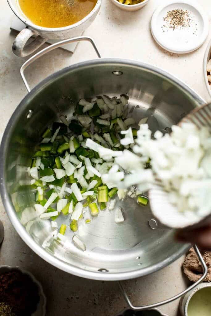 Adding diced onion to a stainless steel pot with diced green peppers.