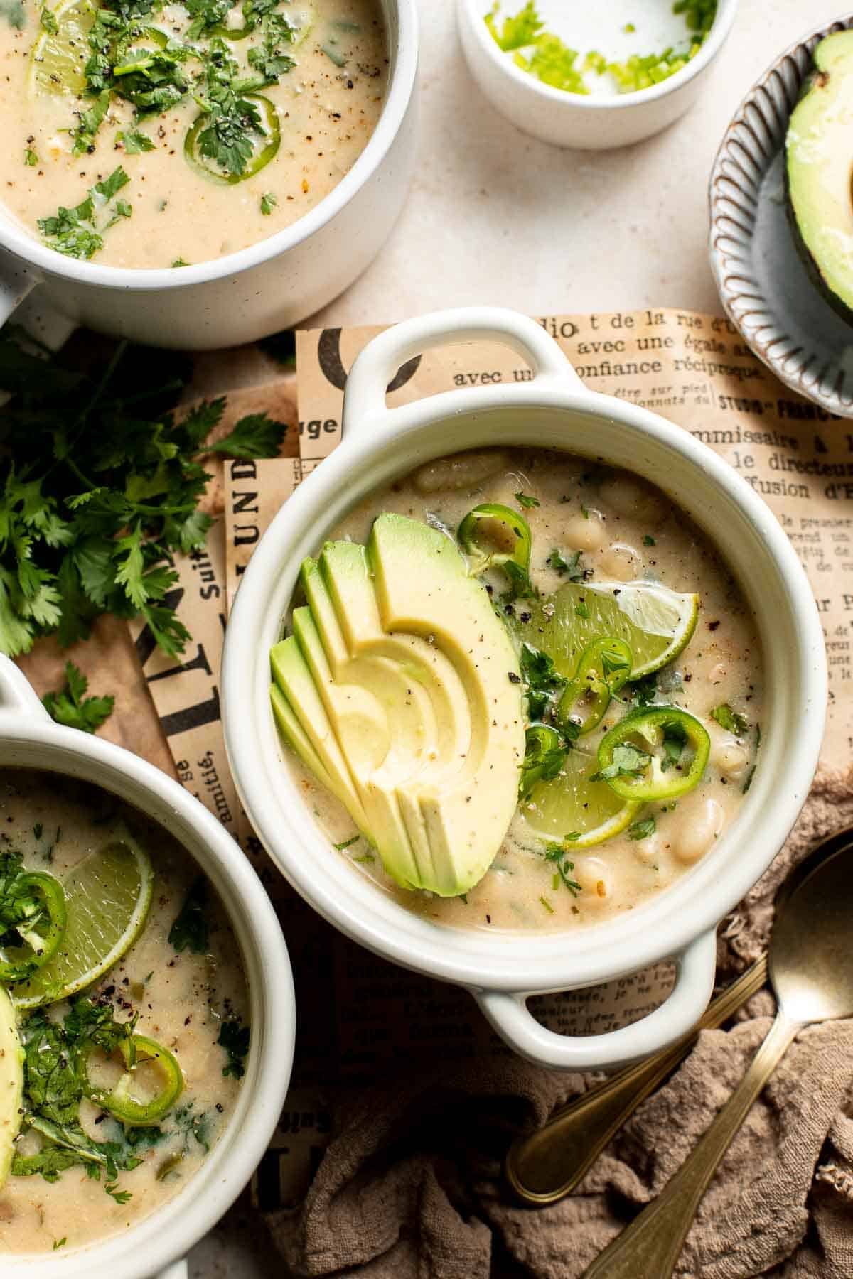 Creamy white bean chili in white bowls topped with fanned avocado slices, lime wedges, sliced jalapeños, and fresh cilantro, on a newspaper-lined board.