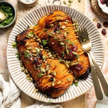 Hasselback butternut squash on a striped platter, topped with dried cranberries, toasted pepitas, and fresh parsley.