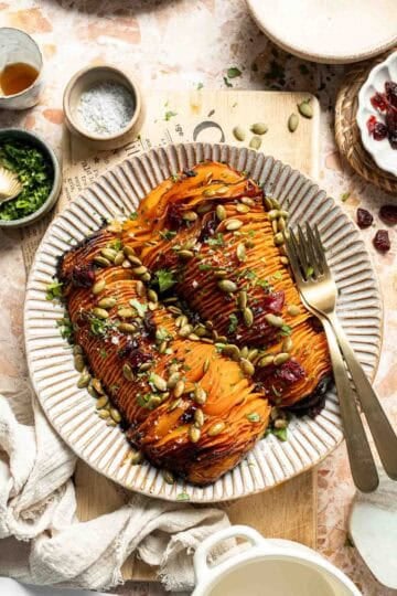 Hasselback butternut squash on a striped platter, topped with dried cranberries, toasted pepitas, and fresh parsley.