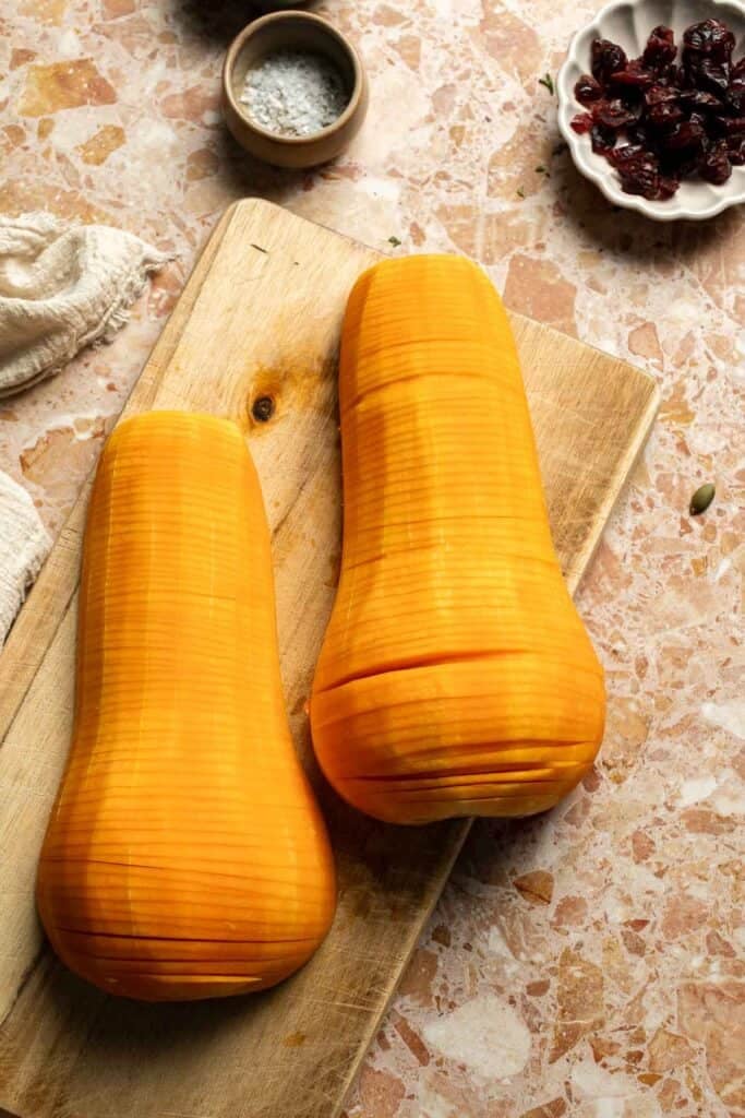 Two butternut squash halves with thin hasselback slices on a cutting board.