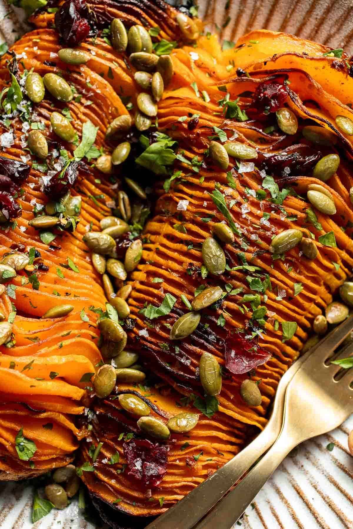 Close-up of hasselback butternut squash with fanned slices, glossy maple glaze, cranberries, pepitas, parsley, and flaky salt.