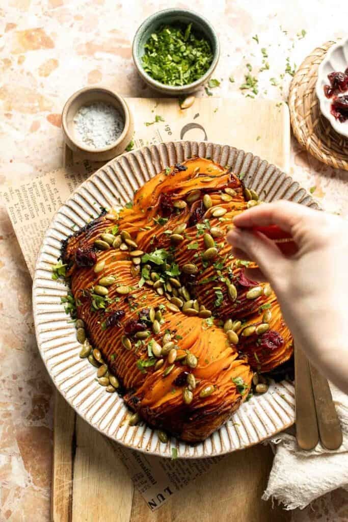 Sprinkling fresh parsley and toppings over roasted hasselback butternut squash on a serving platter.