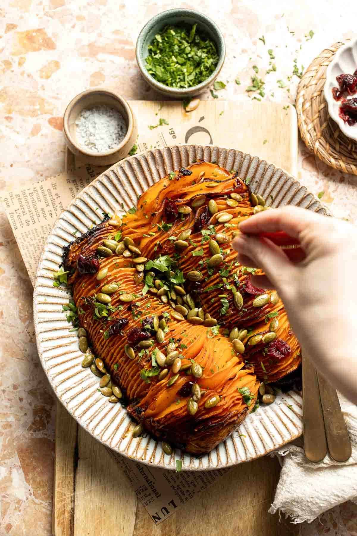 Sprinkling fresh parsley and toppings over roasted hasselback butternut squash on a serving platter.