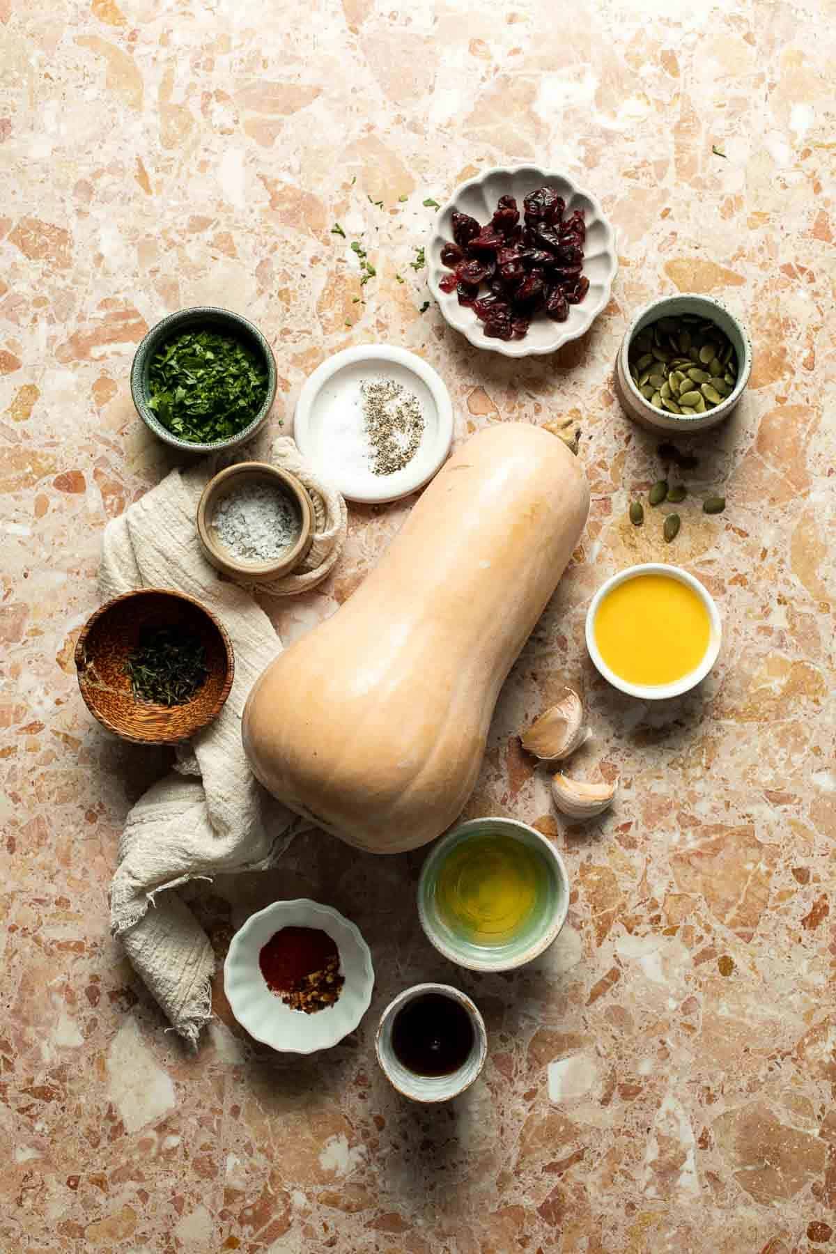 Ingredients for hasselback butternut squash, including a whole butternut squash, olive oil, maple syrup, garlic, thyme, parsley, dried cranberries, spices, and salt, arranged on a pink terrazzo surface.