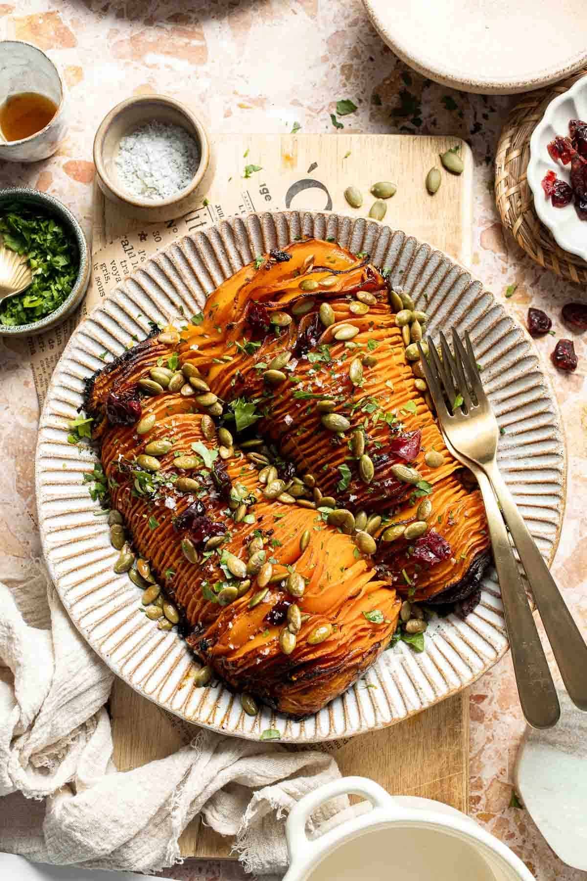 Hasselback butternut squash on a striped platter, topped with dried cranberries, toasted pepitas, and fresh parsley.