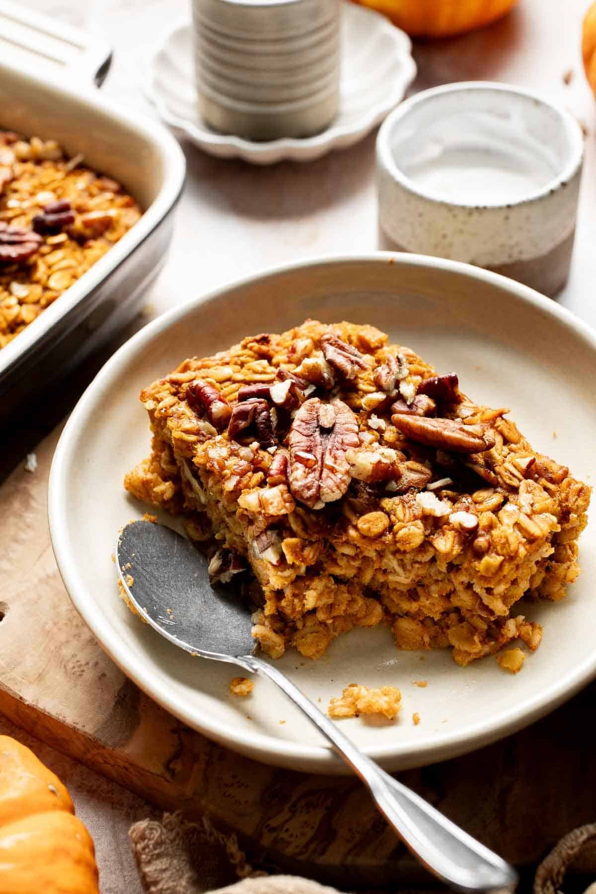 Square of pumpkin baked oatmeal with pecans on a plate with a spoon, next to the baking dish