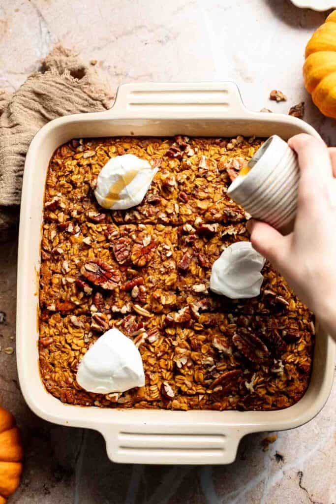 Hand drizzling maple syrup over pumpkin baked oatmeal topped with Greek yogurt