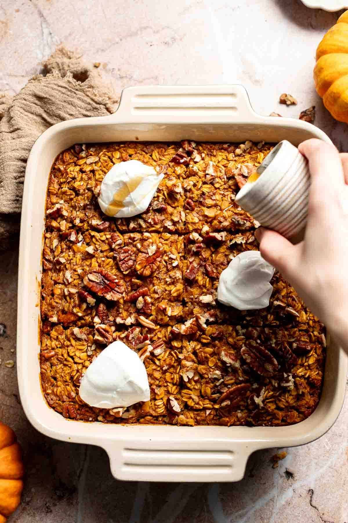 Hand drizzling maple syrup over pumpkin baked oatmeal topped with Greek yogurt