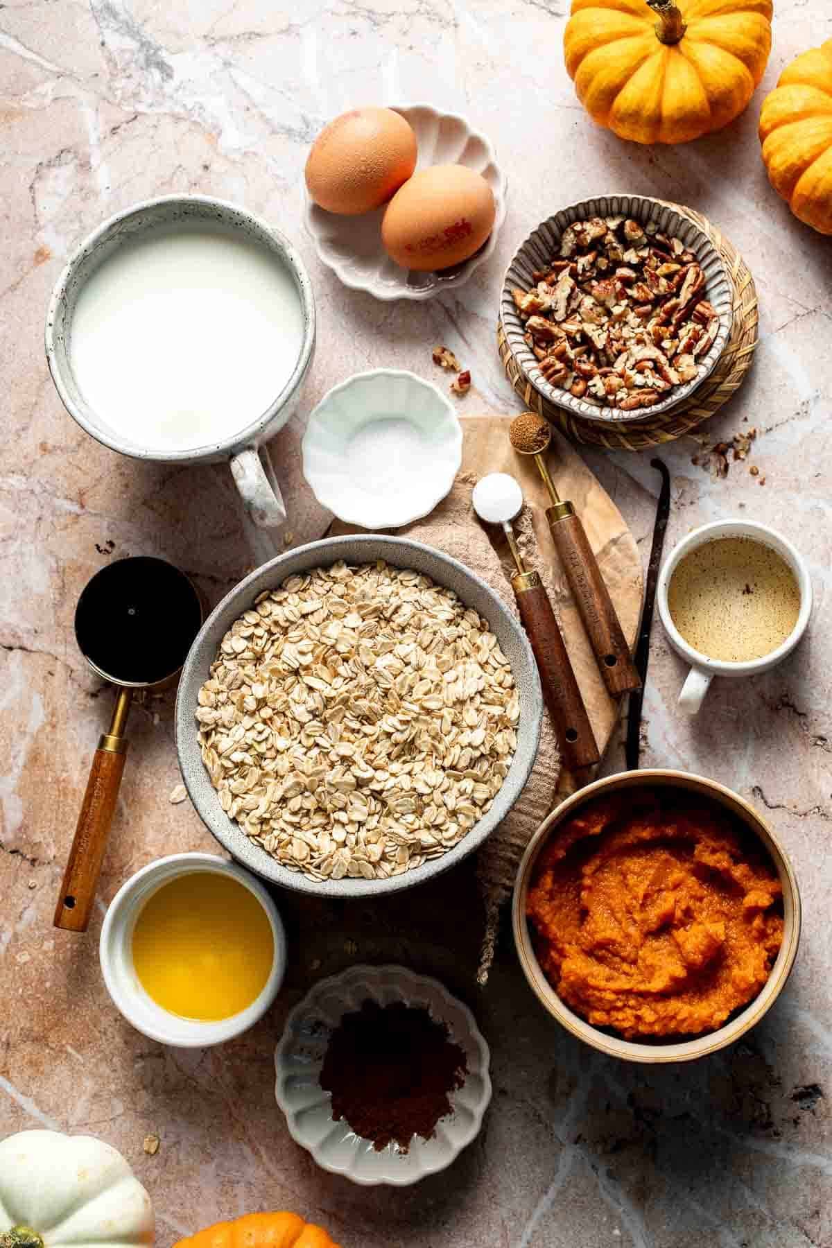Ingredients for pumpkin baked oatmeal with pecans, including rolled oats, pumpkin puree, eggs, milk, maple syrup, and spices, arranged on a marble surface