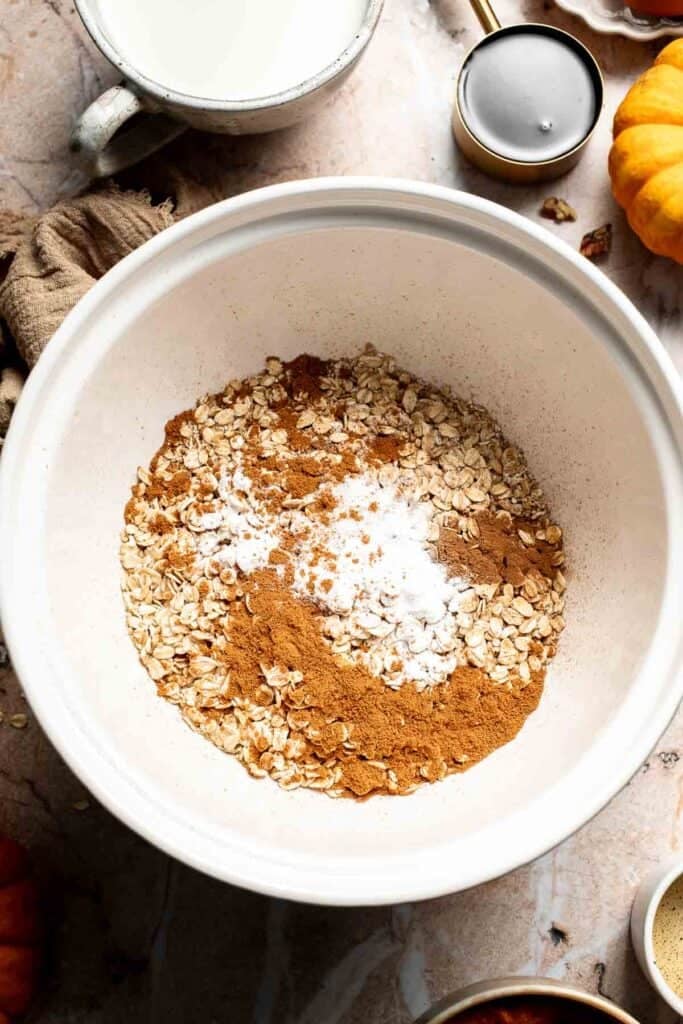 Baking powder, cinnamon, and pumpkin pie spice added on top of rolled oats in a mixing bowl