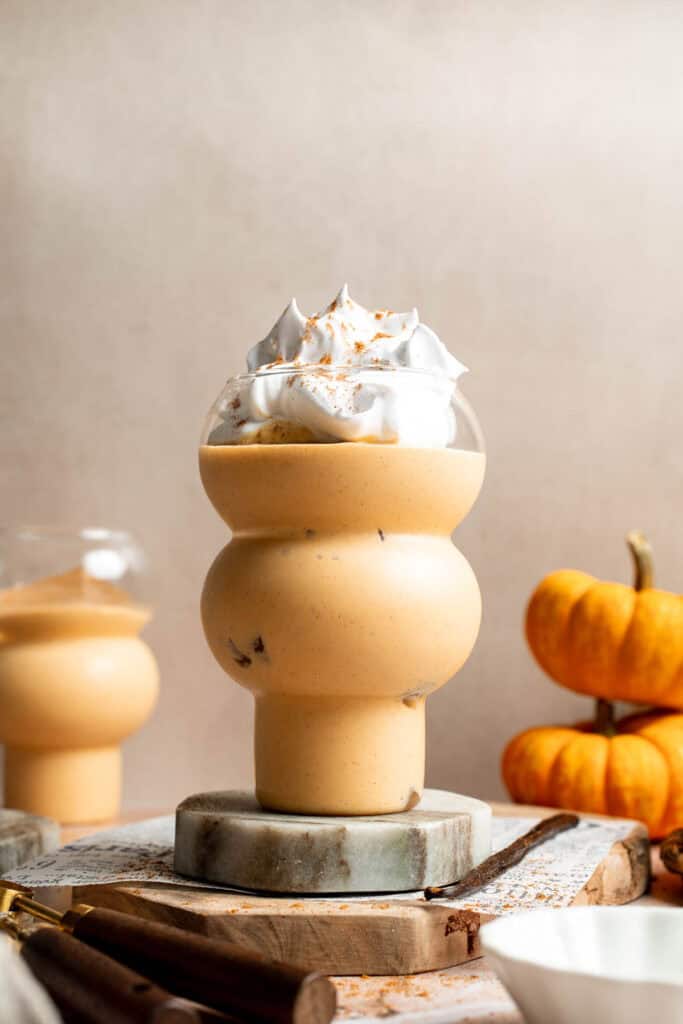 Glass of creamy pumpkin pie smoothie topped with whipped cream and pumpkin spice, with mini pumpkins beside it.