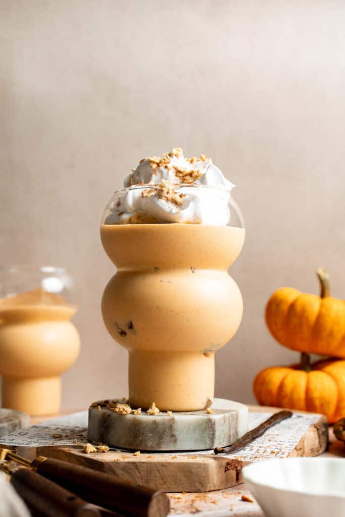 Creamy pumpkin pie smoothie topped with whipped cream and granola in a stacked glass, with mini pumpkins in the background.