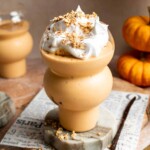 Pumpkin pie smoothie with whipped cream and granola on a marble stand, surrounded by mini pumpkins.