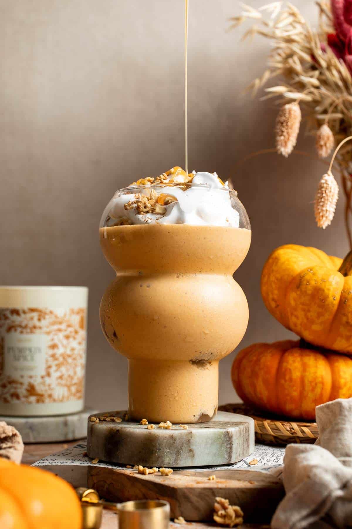 Pumpkin pie smoothie topped with whipped cream and granola with a drizzle of maple syrup, next to a jar of granola and dried grasses.