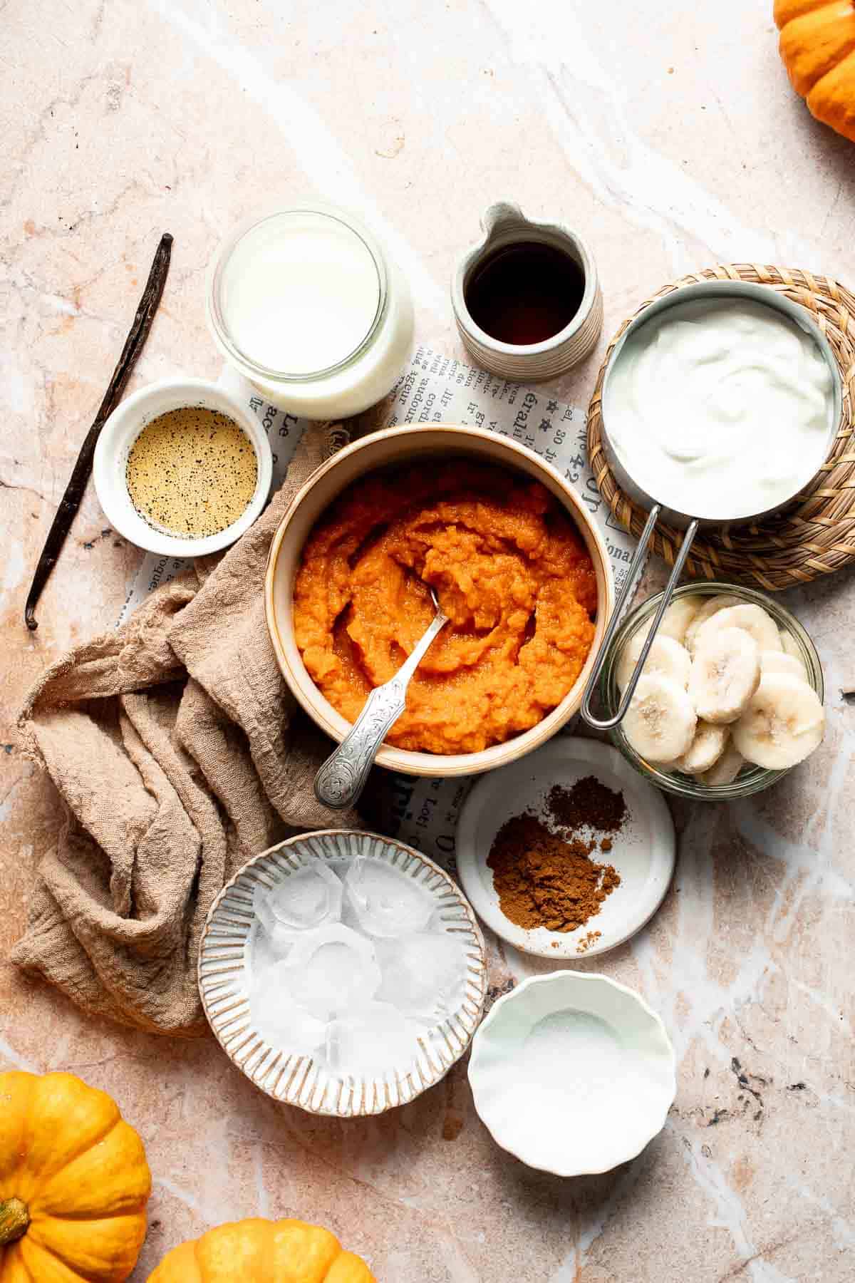 Ingredients for a pumpkin pie smoothie, including pumpkin puree, sliced banana, Greek yogurt, milk, maple syrup, vanilla bean, spices, and ice, laid out on a marble surface with mini pumpkins.