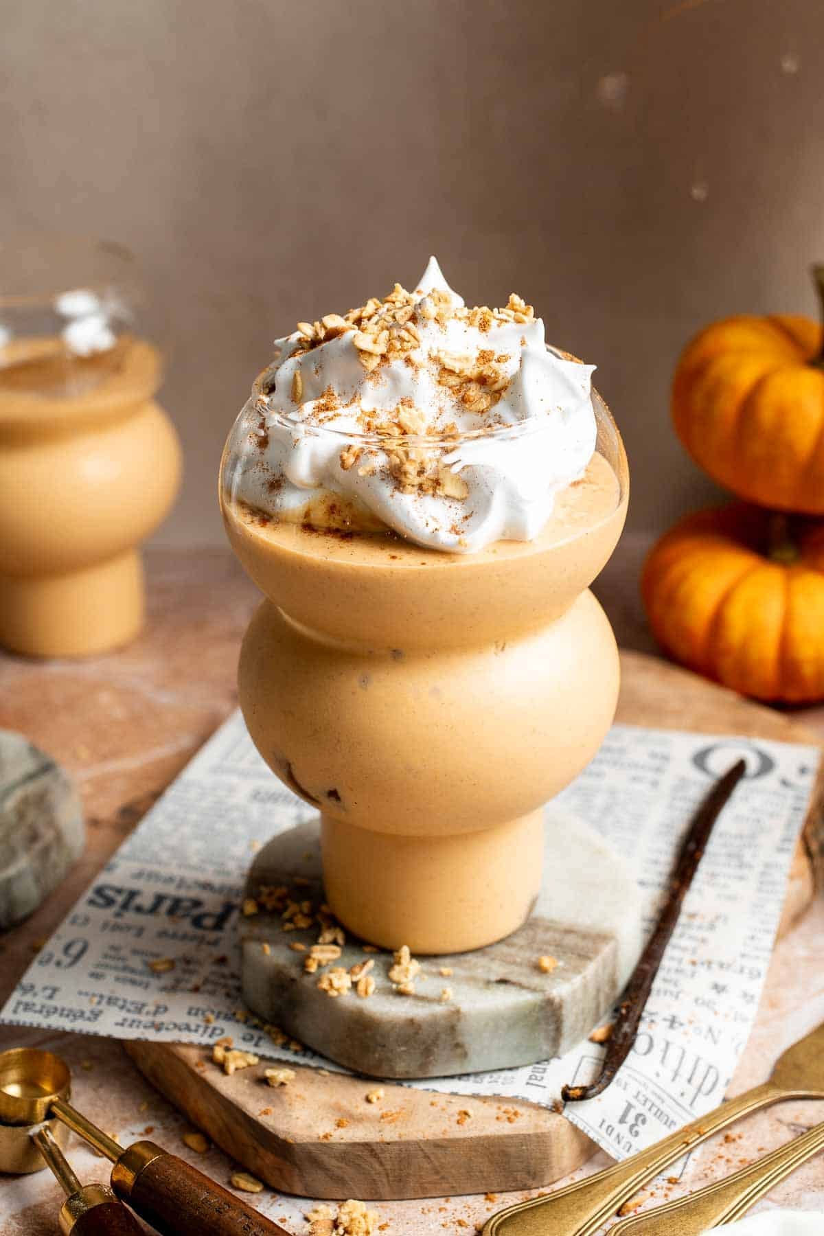 Pumpkin pie smoothie with whipped cream and granola on a marble stand, surrounded by mini pumpkins.