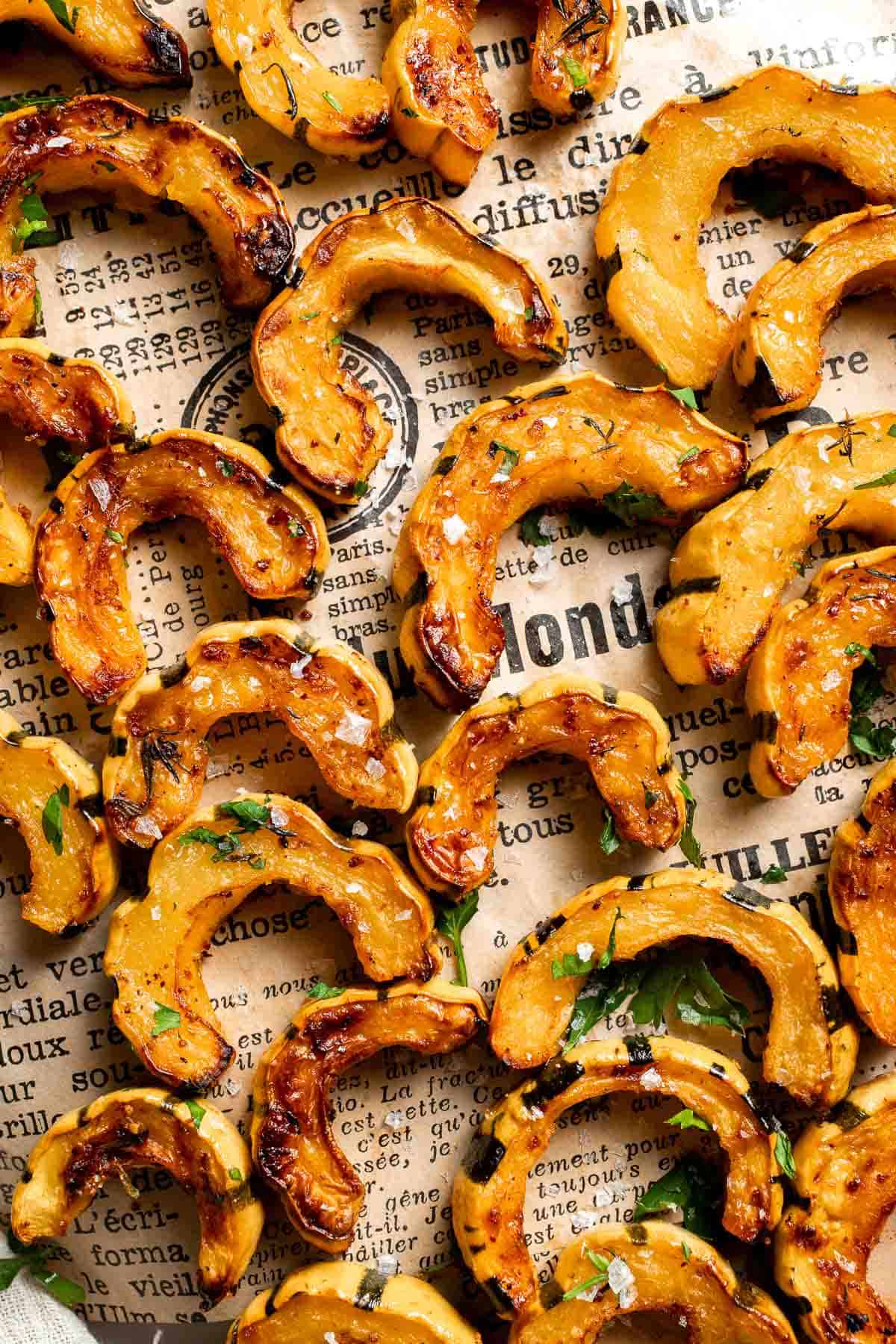 Close-up of roasted delicata squash slices with golden, caramelized edges and flaky salt on a piece of printed paper.