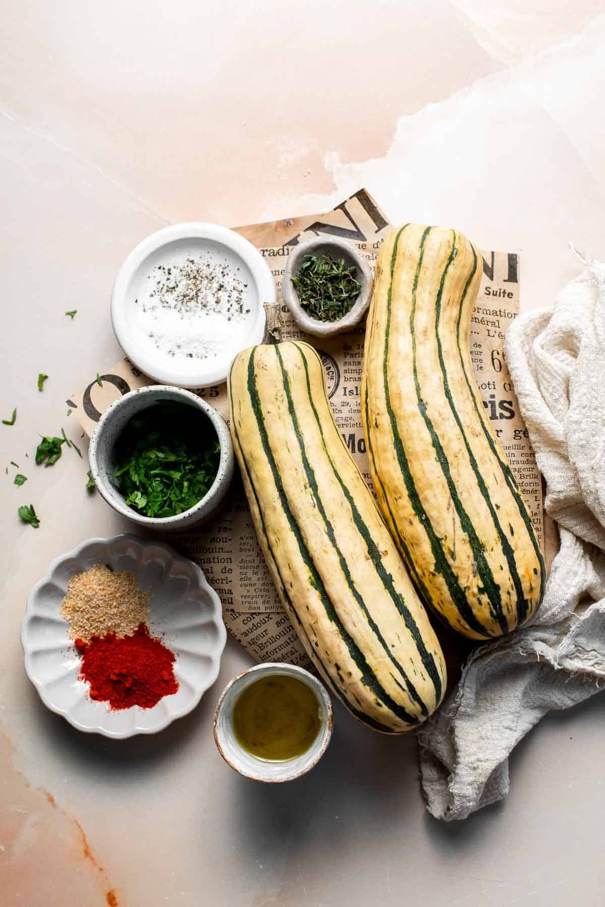 Ingredients for savory roasted delicata squash, including two whole delicata squash, olive oil, garlic powder, smoked paprika, salt, fresh thyme, and parsley, arranged on a light surface.