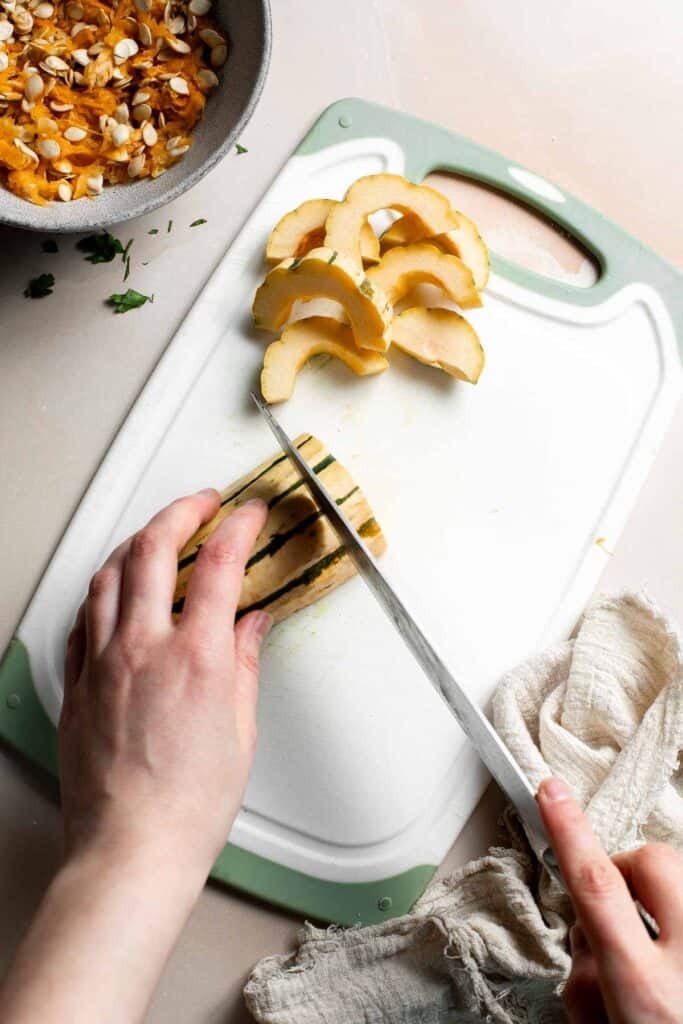 Slicing a delicata squash half into half-moons with a sharp knife on a cutting board.