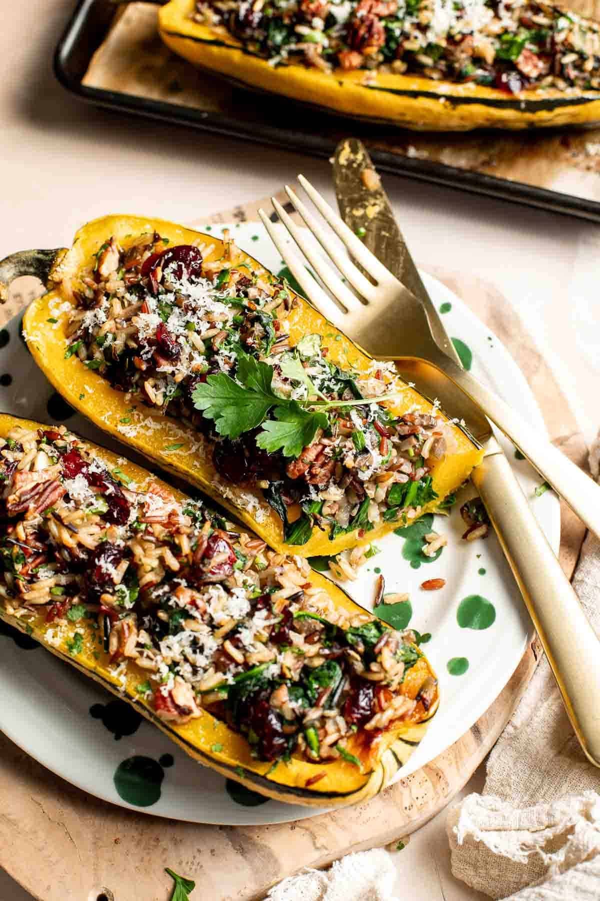 Two stuffed delicata squash halves on a plate with a gold fork and knife, with more squash on the baking sheet behind them.