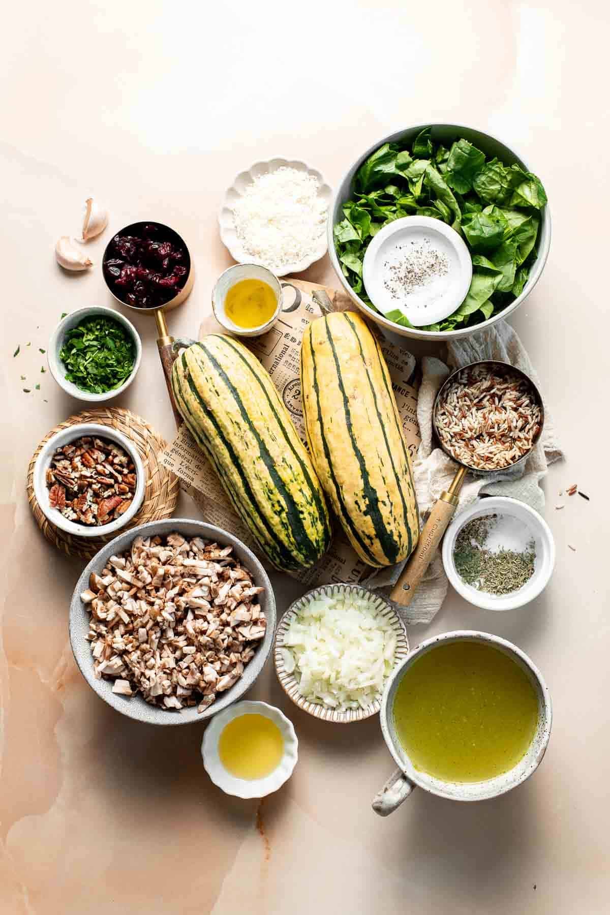 Ingredients for stuffed delicata squash with wild rice, including two whole delicata squash, mushrooms, spinach, wild rice, cranberries, pecans, Parmesan, parsley, and vegetable broth, arranged on a light surface.