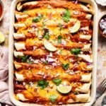 Overhead view of a baking dish of sweet potato black bean enchiladas topped with red onion, cilantro, and lime wedges.