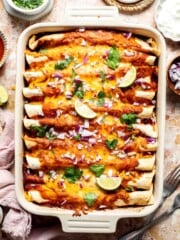 Overhead view of a baking dish of sweet potato black bean enchiladas topped with red onion, cilantro, and lime wedges.