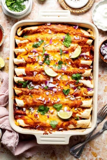 Overhead view of a baking dish of sweet potato black bean enchiladas topped with red onion, cilantro, and lime wedges.